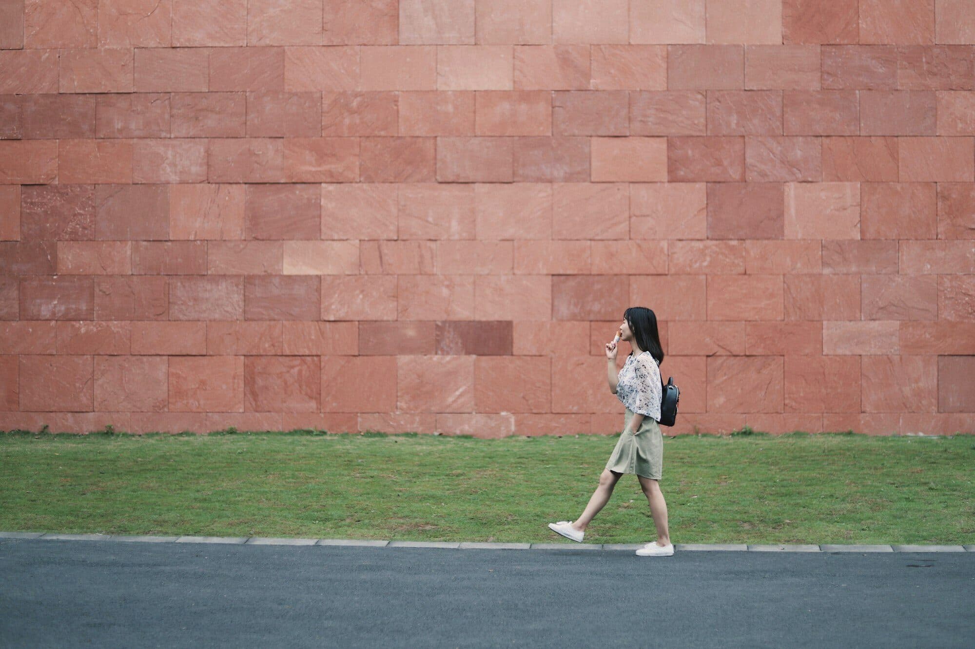 A woman walks briskly past a long terracotta wall on a sunny morning